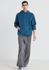 Felpa in pile color turchese con tasca frontale e cordoncino, abbinata a pantaloni gray a gamba larga e scarpe da ginnastica grigie con dettagli bianchi.