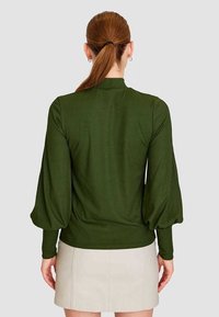 Blusa de canalé verde oscuro con mangas abullonadas, cuello alto y cintura ajustada. Llevada con una falda beige clara, vista desde la parte posterior.