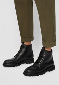Bottines en cuir noir avec des bouts ronds, des semelles en caoutchouc crantées et des œillets en métal, accompagnées de pantalons vert olive retroussés au niveau des chevilles.