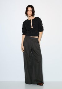 Vrouw staat tegen een witte muur, draagt een zwart, kort, pluizig vest, wijdvallende donkere broek en bruine schoenen, met één hand in de zak.