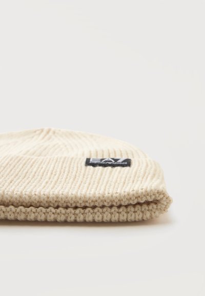 Bonnet en tricot côtelé de couleur crème avec bord replié et petite étiquette noire à l’avant sur un fond clair uni.