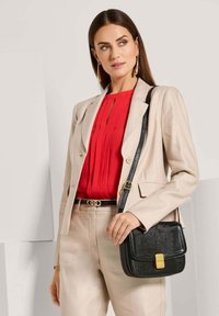 Zwarte leren schoudertas met een textuurafwerking, gouden sluiting en verstelbare schouderband. Gemaakt in combinatie met een beige blazer en een rode geplooide top.