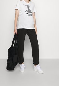 Valkoinen t-paita, jossa on musta nuudelikulho -grafiikka ja teksti "I ❤️ NOODLES", yhdistettynä löysiin mustiin housuihin ja valkoisiin lenkkareihin. Mukana musta kassi.