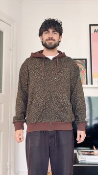 Νέος άνδρας με σκούρα σγουρά μαλλιά και γένια, φορώντας καφέ φούτερ με λεοπάρ σχέδιο και σκουρόχρωμο παντελόνι, στέκεται μέσα σε εσωτερικό χώρο κοντά σε τηλεόραση και κορνιζαρισμένη τέχνη.