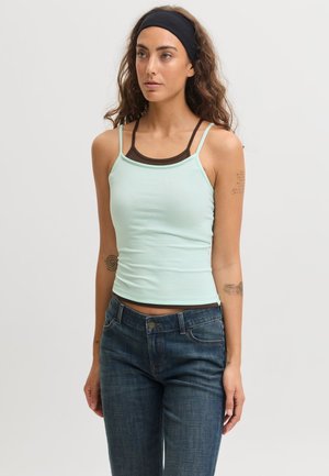 Femme aux longs cheveux bouclés avec un bandeau noir, portant un caraco bleu clair par-dessus un haut marron et un jean bleu sur un fond uni.