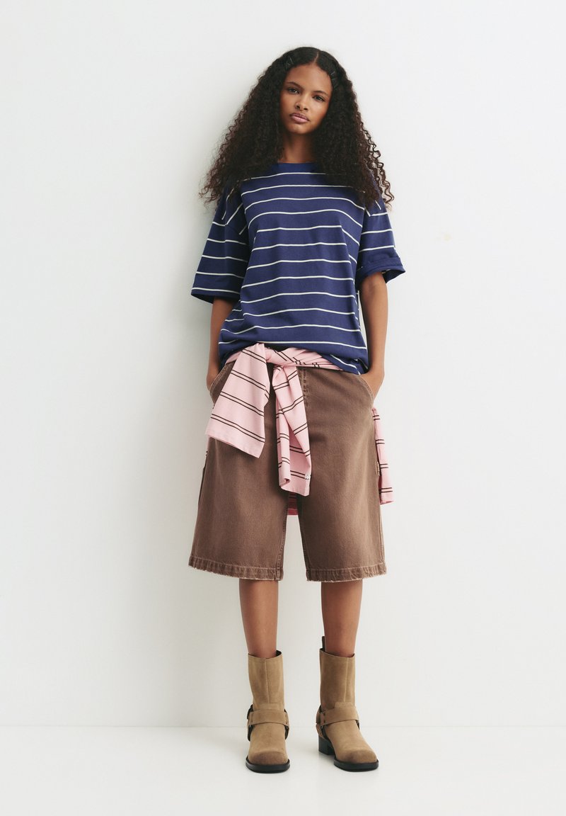 Marineblå stribet t-shirt, afslappet pasform, parret med brune denim bermuda shorts. Lys pink skjorte bundet om taljen, som fuldender outfittet med sandfarvede støvler.