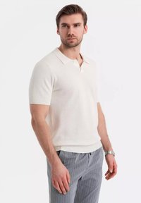 Polo triko s krátkým rukávem v krémové barvě s texturovaným pletením, s klasickým límečkem a žebrovanými manžetami, v kombinaci se šedými kalhotami s jemným proužkem.