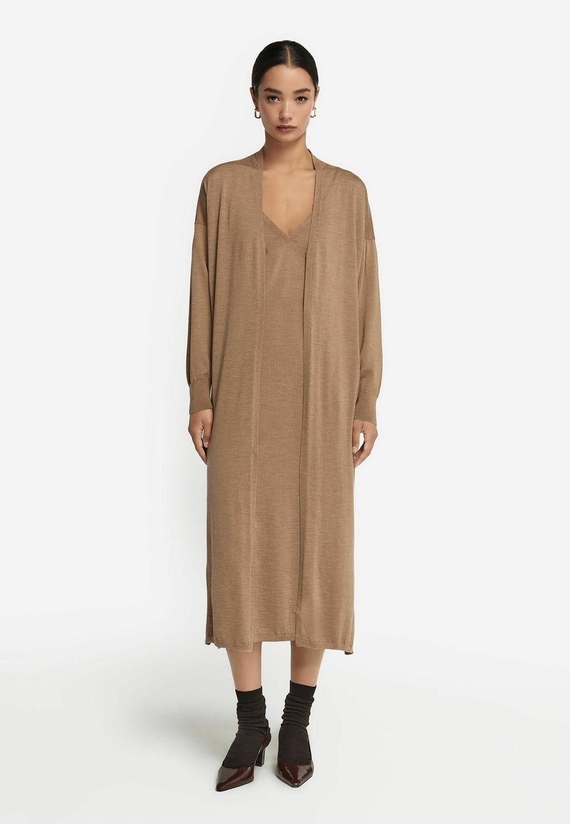 Lång, brun kofta klänning med V-ringning, långa ärmar och rak passform. Mjuk struktur, sidan slits, kombinerad med mörka klackskor och strumpor.