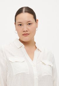 Chemise blanche à boutons avec des rayures verticales dorées, comportant deux poches poitrine, un col et une texture légère et douce.