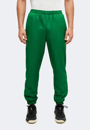 Homme portant un pantalon de jogging vert vif à taille et chevilles élastiques, associé à des chaussures de sport noires sur un fond clair uni.