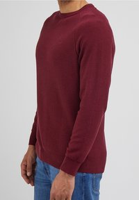 Pull en maille bordeaux avec col rond, poignets et ourlet côtelés. Motif texturé sur l'ensemble, coupe ajustée, porté avec un jean bleu.