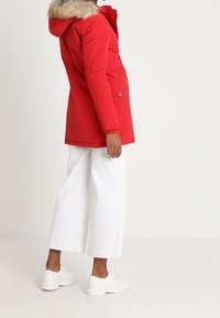 Rote Parkajacke mit fellgefütterter Kapuze, Reißverschluss- und Knopfverschluss, seitlichen Taschen, kombiniert mit weißen weit geschnittenen Hosen und weißen Sneakers.