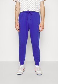 Lila joggingbyxor i mjukt material, med dragsko i midjan och smalnande manschetter. En liten logotyp syns på vänster sida.