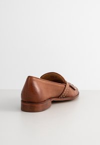 Chaussure plate en cuir marron avec un petit talon, ornée de clous décoratifs sur le côté. Texture lisse et design à bout rond.