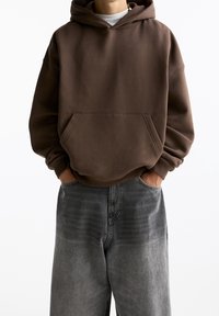 Sweat à capuche marron avec une poche avant, associé à un jean gris ample. Le tissu a une texture douce, et la coupe est décontractée.
