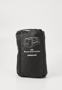 Crna Patagonia 40L Black Hole torba za odlaganje pakovana unutar svog kompaktnog crnog spremnika s patentnim zatvaračem i ručkom na običnoj pozadini.