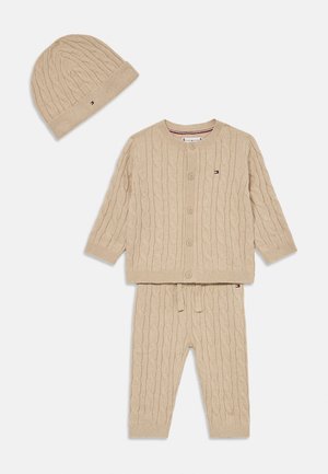 Conjunto de bebé em malha canelada beige, com cardigan abotoado, calças com cordão e gorro combinando, todos com pequenos patches do logótipo da marca.