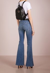 Sac à dos en cuir noir avec détail logo, porté par-dessus un t-shirt blanc. Modèle en jean évasé bleu et talons noirs, vu de dos.