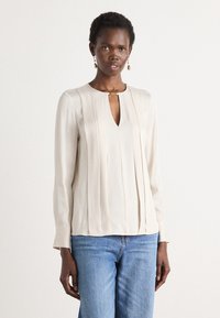 Lys beige langermet bluse med vertikale plisseringer, V-formet halsutskjæring og en gullfarget detalj ved kragen. Kombinert med blå jeans.