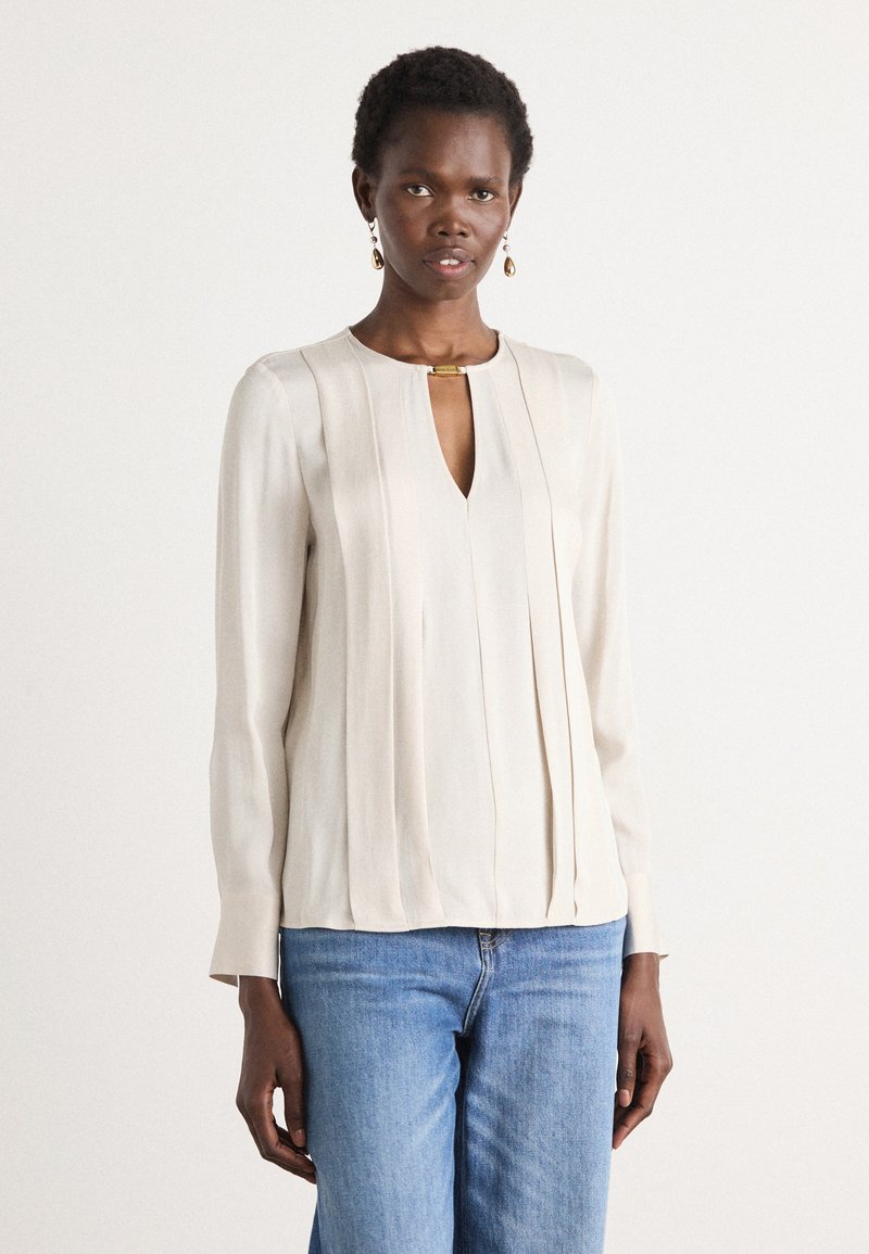Lys beige langermet bluse med vertikale plisseringer, V-formet halsutskjæring og en gullfarget detalj ved kragen. Kombinert med blå jeans.