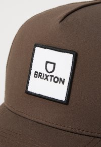 Barna, texturált anyagból készült sapka, melyen egy fehér négyzet alakú folt található fekete betűkkel, melyen az olvasható, hogy "BRIXTON", valamint egy pajzs logo.