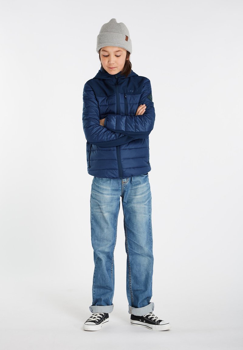 Mørkeblå pufferjakke med hætte, lynlås foran og brystlomme. Matcher med lyseblå jeans og sorte sneakers. Grå strikket hue på hovedet.