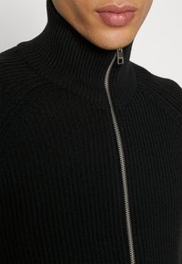 Svart ribbad stickad tröja med hög krage och silverdetalj i form av dragkedja framtill. Tätsittande och strukturert tyg.