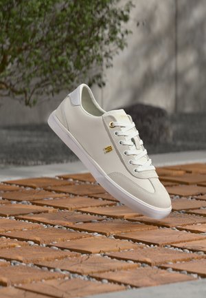 Λευκό sneaker χαμηλού τύπου με χρυσό λογότυπο, λευκά κορδόνια και σουέτ λεπτομέρειες, αιωρούμενο πάνω από ένα ξύλινο τετράγωνο και έδαφος με μοτίβο από βότσαλα σε εξωτερικό χώρο.
