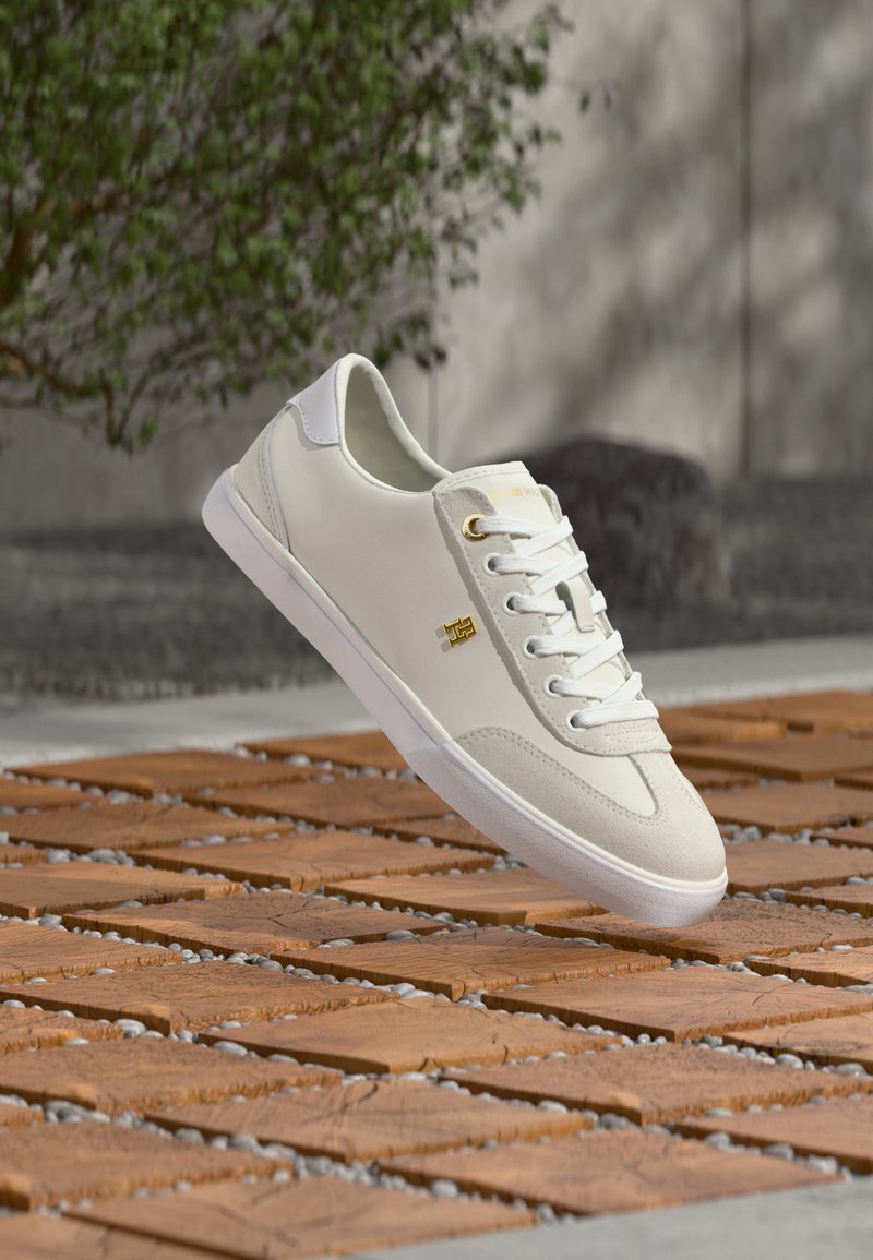 Λευκό sneaker χαμηλού τύπου με χρυσό λογότυπο, λευκά κορδόνια και σουέτ λεπτομέρειες, αιωρούμενο πάνω από ένα ξύλινο τετράγωνο και έδαφος με μοτίβο από βότσαλα σε εξωτερικό χώρο.