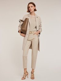 Beżowy długi płaszcz na lekkim sweterku i beżowych spodniach wysokim stanem. Brązowa torebka o eleganckim designie. Brązowe sandały na obcasie z wzorzystym paskiem.