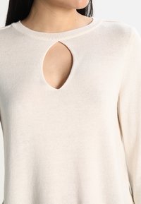 Sweter w kolorze kremowym, dziany, z okrągłym dekoltem i wycięciem w kształcie łezki. Miękka struktura, długie rękawy oraz luźny krój.
