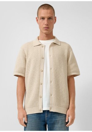 Mężczyzna o krótkich włosach, ubrany w beżową, teksturowaną koszulę na krótki rękaw, zapinaną na guziki, na białą koszulkę i niebieskie dżinsy, stojący na jednolitym tle.