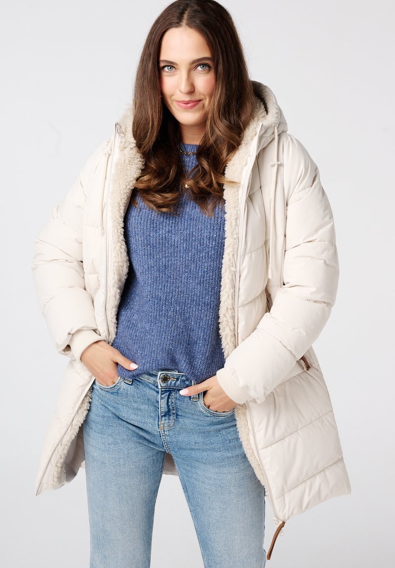 Pufferjack in lichtbeige met een nepbontvoering, capuchon en ribgebreide manchetten, gecombineerd met een blauwe textuurtrui en lichtdenim spijkerbroek.