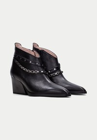 Botas de tobillo de cuero negro con puntas afiladas y tacones de cuña. Presentan un detalle de cadena y una correa con tachuelas. Acabado texturizado con un forro suave.