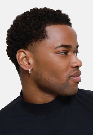 Boucles d'oreilles en argent en forme de créoles avec une finition brossée, portées à l'oreille gauche. La personne porte un haut à col roulé noir, profil latéral visible.