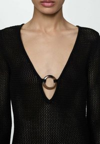 Top negru tricotat, cu un decolteu adânc în formă de V și un accent oval din metal la centru. Țesătura are o textură ușoară, cu o țesătură deschisă.