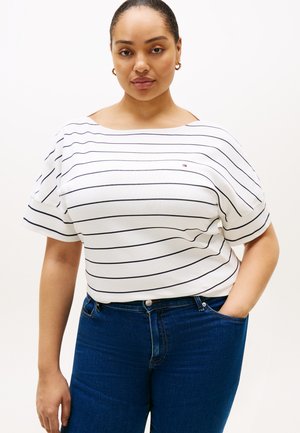 Mujer que lleva una camiseta blanca de manga corta con rayas horizontales negras y vaqueros azules, de pie con una mano en el bolsillo contra un fondo liso.