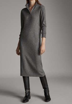 Femme portant une longue robe en maille grise avec un col zippé et des bottes en cuir noires jusqu'aux genoux, debout devant un fond gris uni.