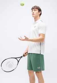 Hvit poloskjorte med svarte detaljer, kombinert med grønne shorts. Personen holder et svart tennisracket. En gul tennisball er i luften.