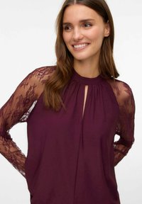Blusa bordeaux con maniche in pizzo trasparente, scollatura alta, dettaglio a chiave e vestibilità morbida. Tessuto morbido con accenti di pizzo floreale sulle maniche.