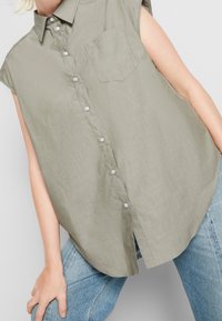 Camicia verde oliva senza maniche con bottoni, realizzata in tessuto leggero e lavorato. Presenta una tasca frontale e un orlo curvo. Abbinata a jeans chiari.