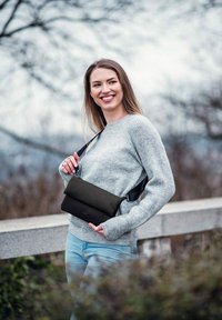 Černá crossbody taška vyrobená z měkkého materiálu, skládací design, nastavitelný popruh, nošena přes šedý svetr a modré džíny. Přírodní venkovní prostředí.