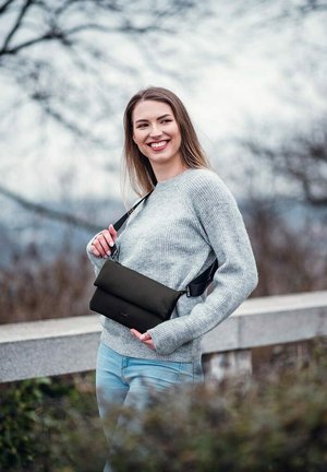 Černá crossbody taška vyrobená z měkkého materiálu, skládací design, nastavitelný popruh, nošena přes šedý svetr a modré džíny. Přírodní venkovní prostředí.