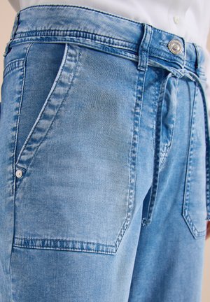 Gros plan sur un jean en denim bleu clair avec des poches plaquées à l’avant, des passants de ceinture, un bouton et une taille à cordon noué, porté par une personne vêtue d’une chemise blanche.