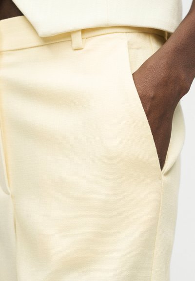 Σκούρο χέρι που ακουμπάει μέσα στην τσέπη παντελονιού κρεμ χρώματος με ραμμένη εφαρμογή, συνοδευόμενο από ταιριαστό θηλάκι ζώνης και μέση.