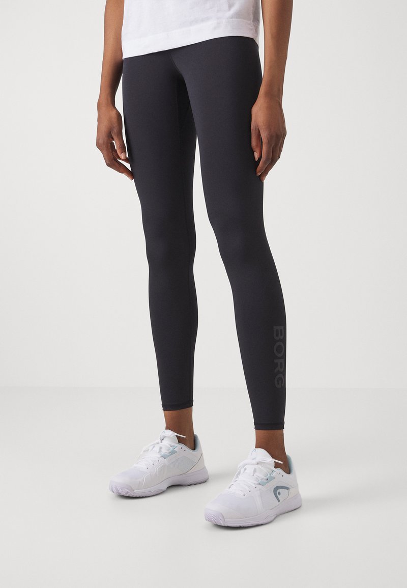 Leggings pretos justos com uma textura suave e um logótipo visível ao longo do lado, combinados com sapatilhas brancas desportivas com detalhes em azul claro.