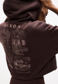 Άτομο που φοράει σκούρο φούτερ με κουκούλα, με εξοπλισμό γυμναστικής και τέσσερις αθλητές τυπωμένους στην πλάτη, σηκώνοντας το χέρι κοντά στο κεφάλι.