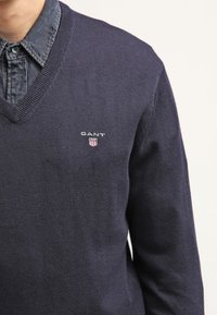 Granatowy sweter dziany z dekoltem w serek, długimi rękawami oraz małym logo GANT w kształcie tarczy po lewej stronie klatki piersiowej.