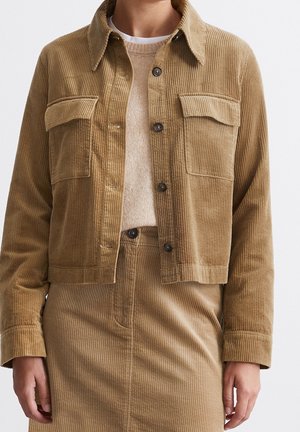 Persoon draagt een lichtbruine corduroy jas en bijpassende rok met een beige trui, zichtbaar van nek tot heupen.