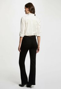 Camicia bianca a maniche lunghe con colletto arricciato, abbinata a jeans neri a vita alta e svasati. Il modello indossa tacchi neri, con le braccia rilassate.